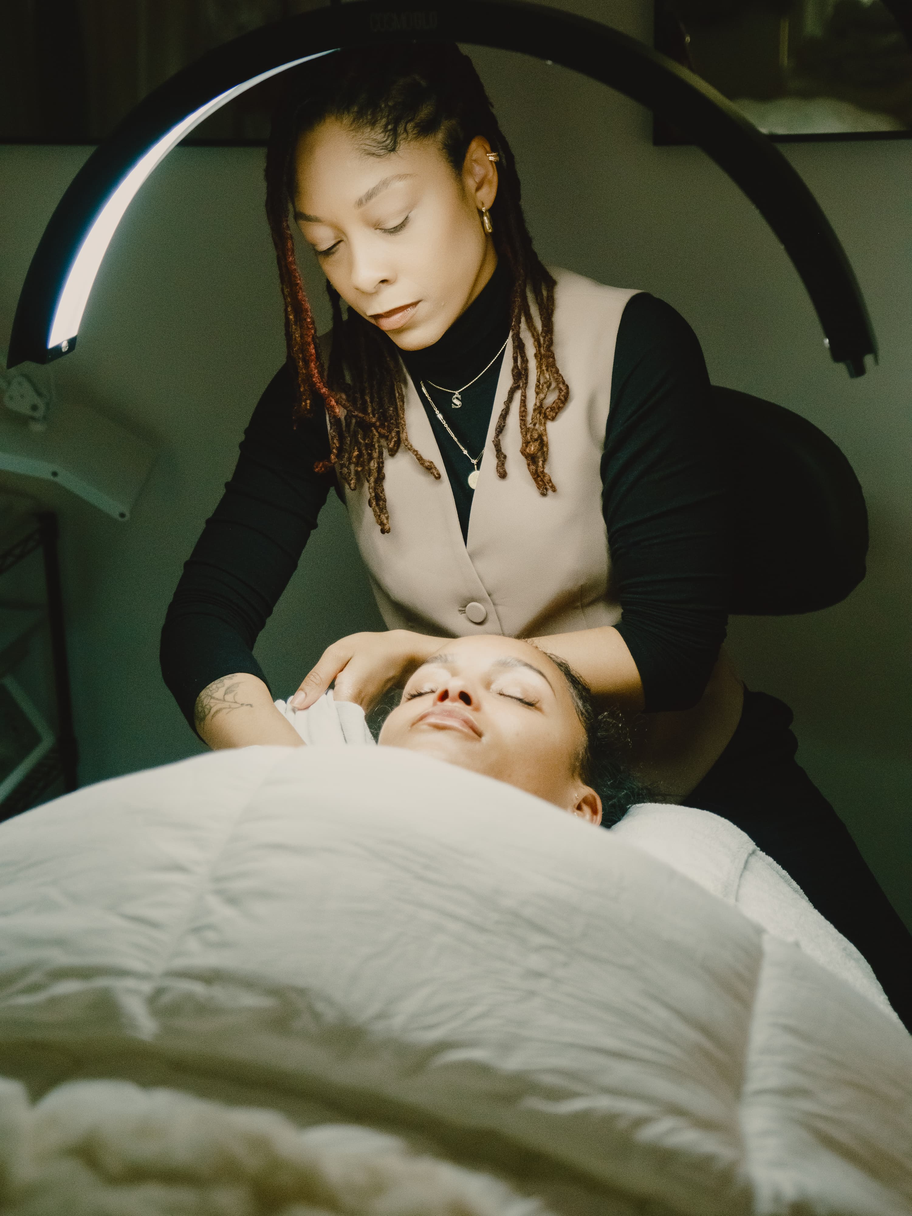 Esthetician in a tan vest and black turtleneck giving a facial to a client lying under a white blanket beneath an arched light
