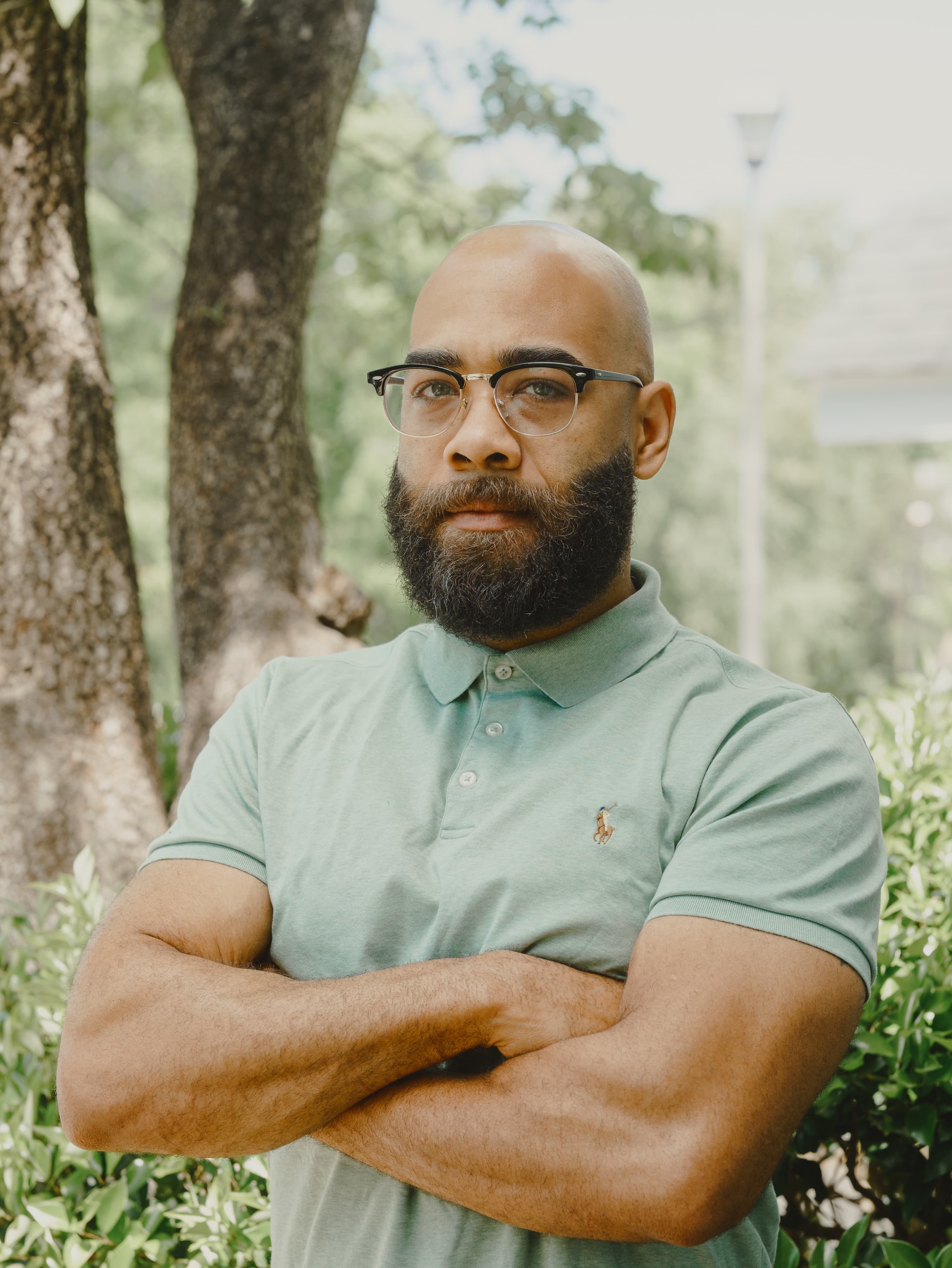 Man with a shaved head, beard and glasses in a sage green polo standing with arms crossed in front of a tree and greenery