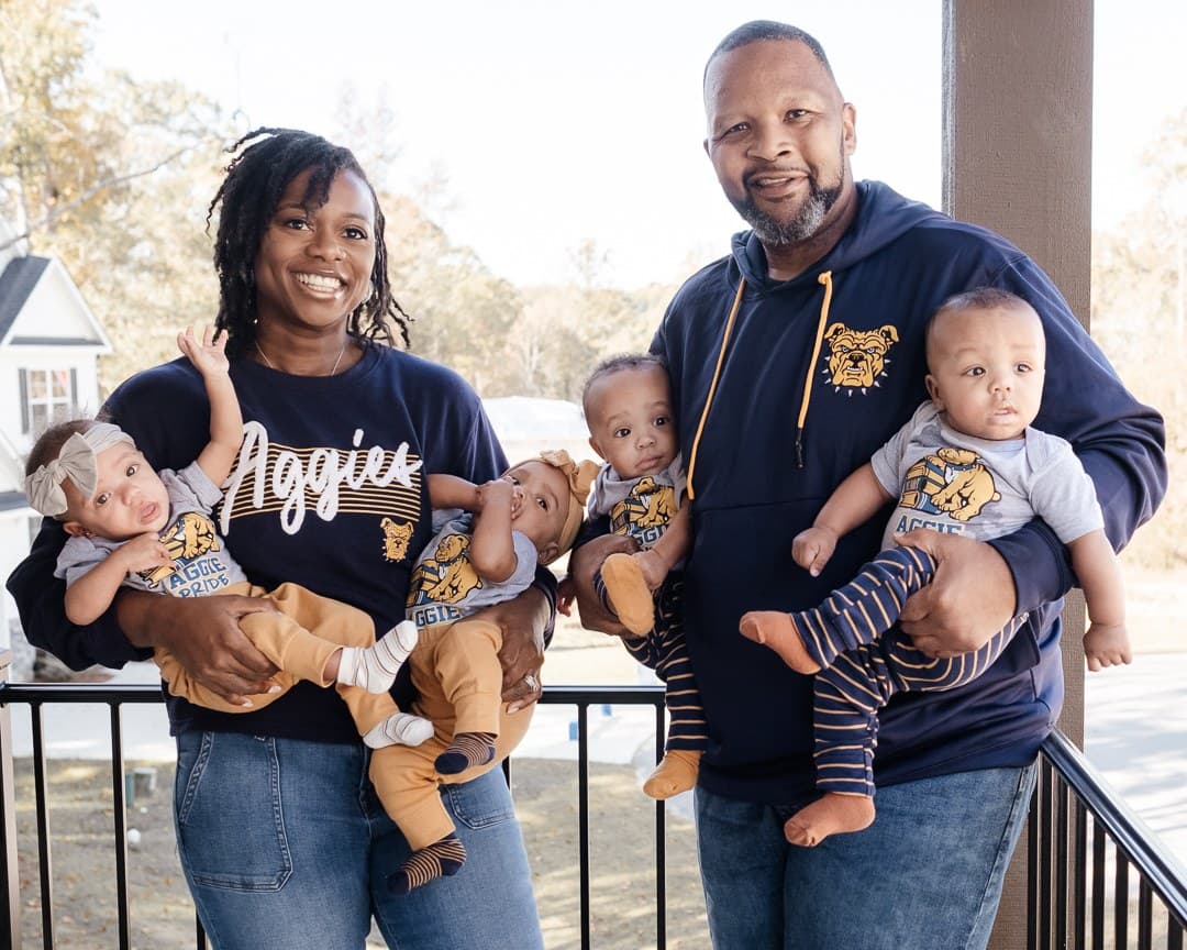 Parents in matching Aggies shirts holding their four babies on a porch