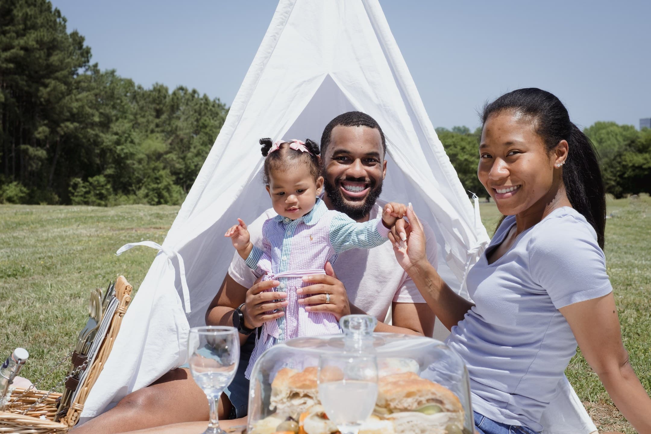 Parents and their toddler daughter smiling at a picnic in front of a white teepee