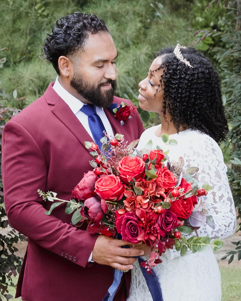 Groom in a burgundy suit and bride in lace smiling at each other over a red bouquet