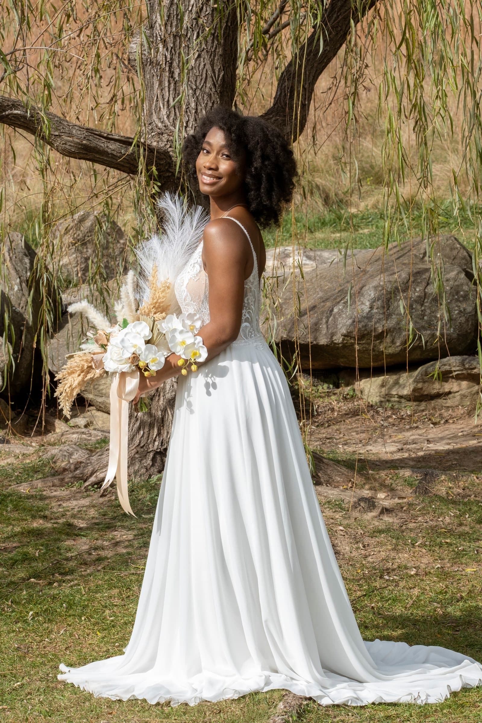 Bride smiling over her shoulder with a pampas and orchid bouquet under a willow tree