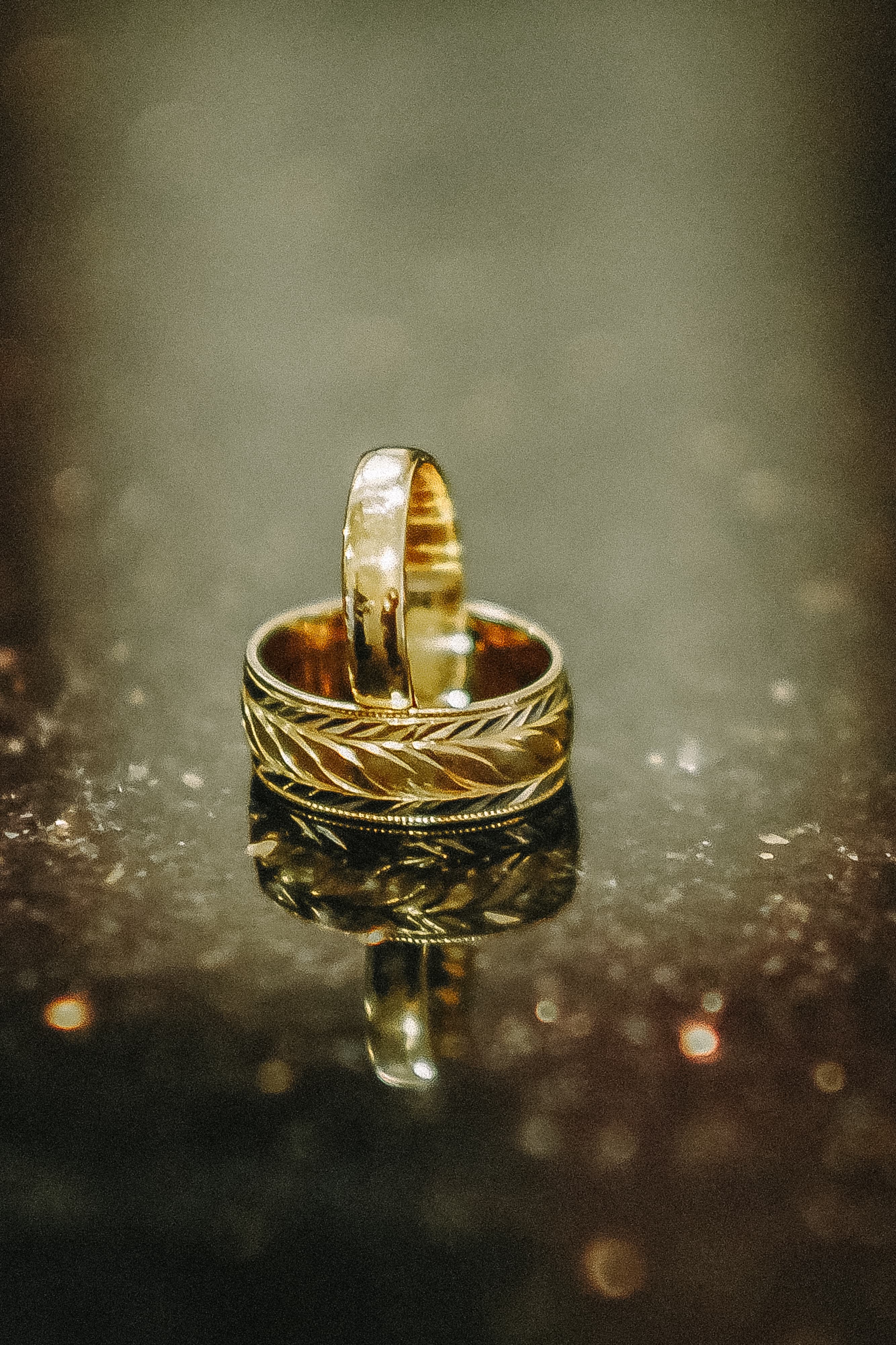 Two gold wedding bands, one standing upright inside an engraved band, reflected on a dark glittering surface