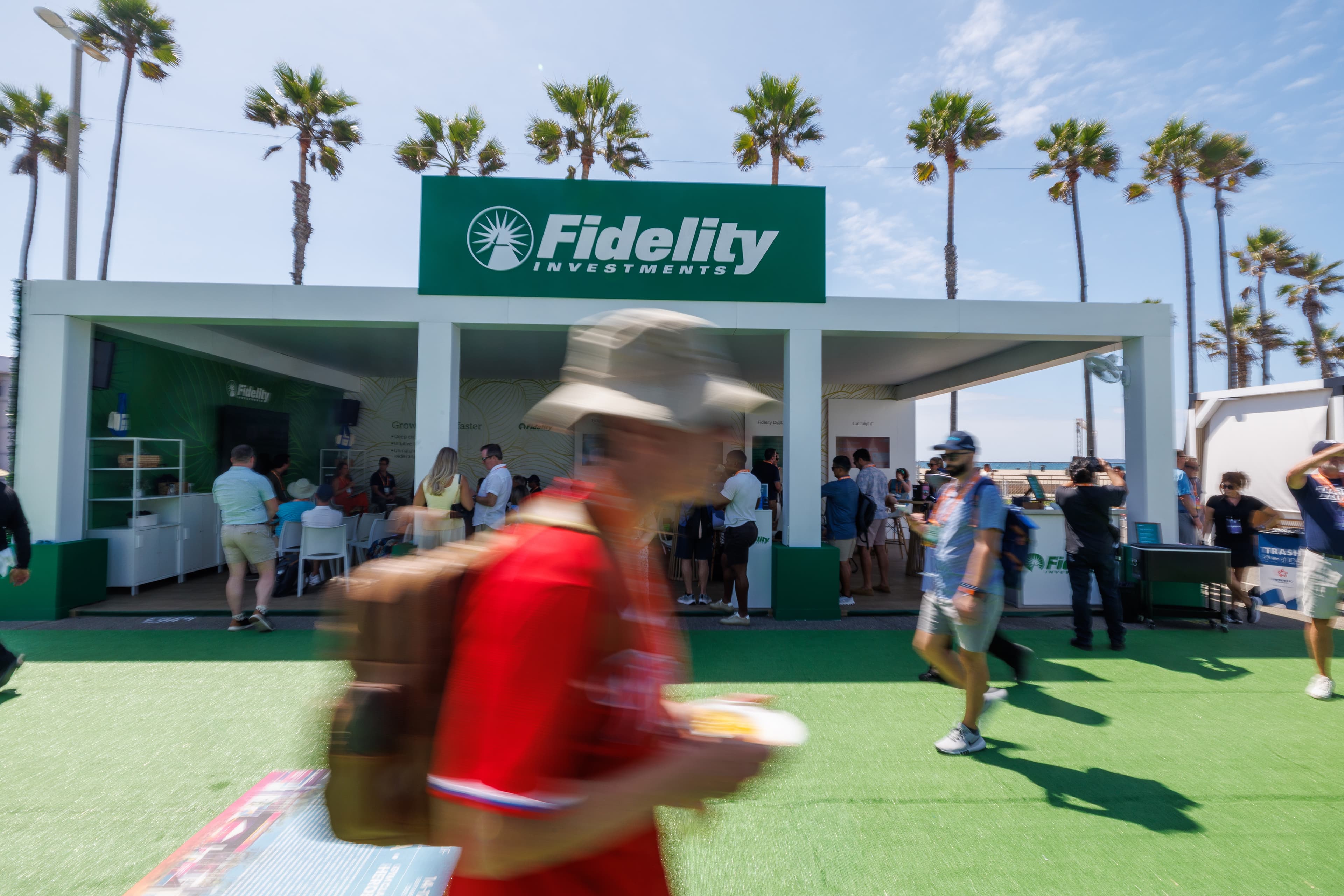 Attendees gathered at a sponsor booth beneath palm trees as a guest walks past at an outdoor conference