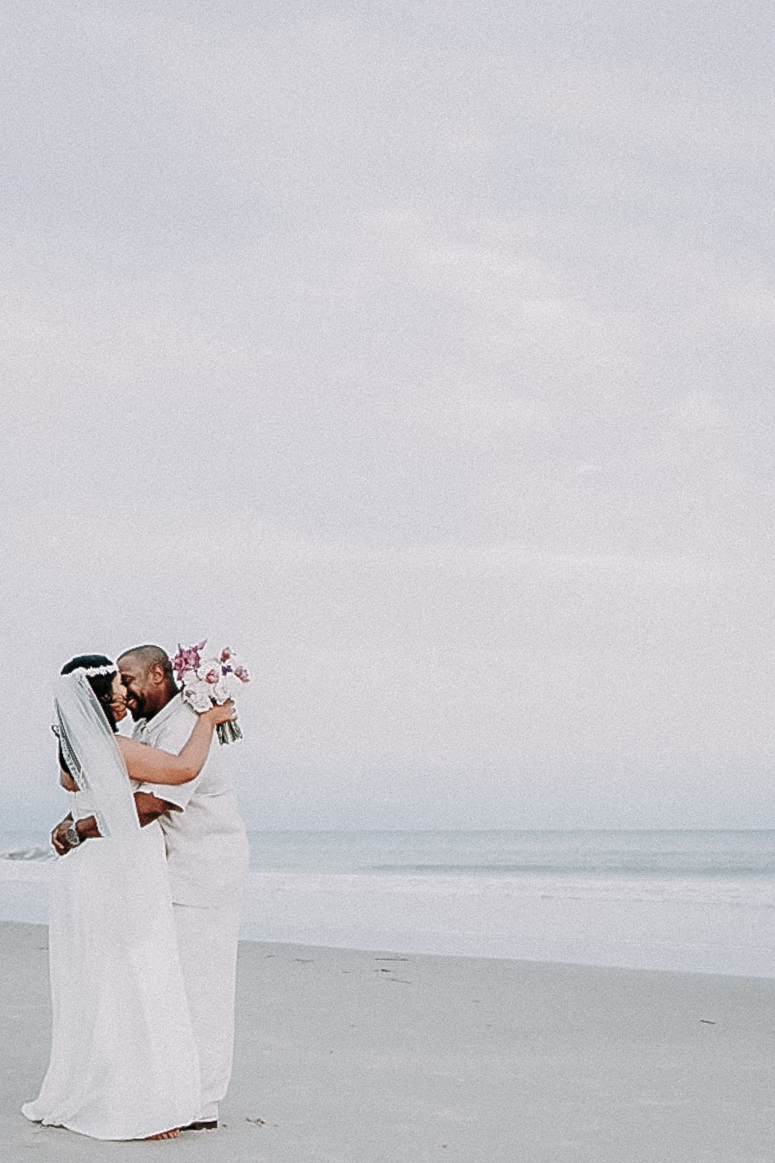 Couple in white embracing on a beach under an overcast sky, with the bride holding a pink and white bouquet