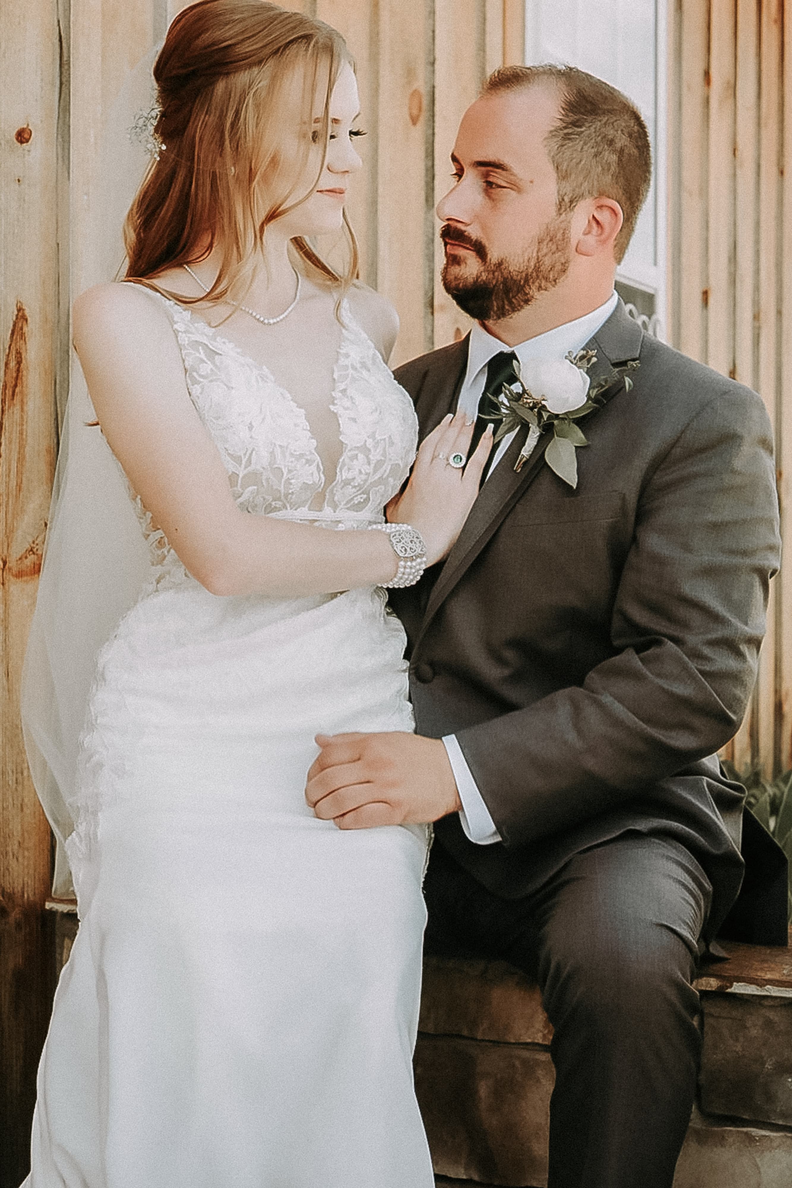 Bride in a lace gown seated beside a groom in a gray suit against a wood plank wall