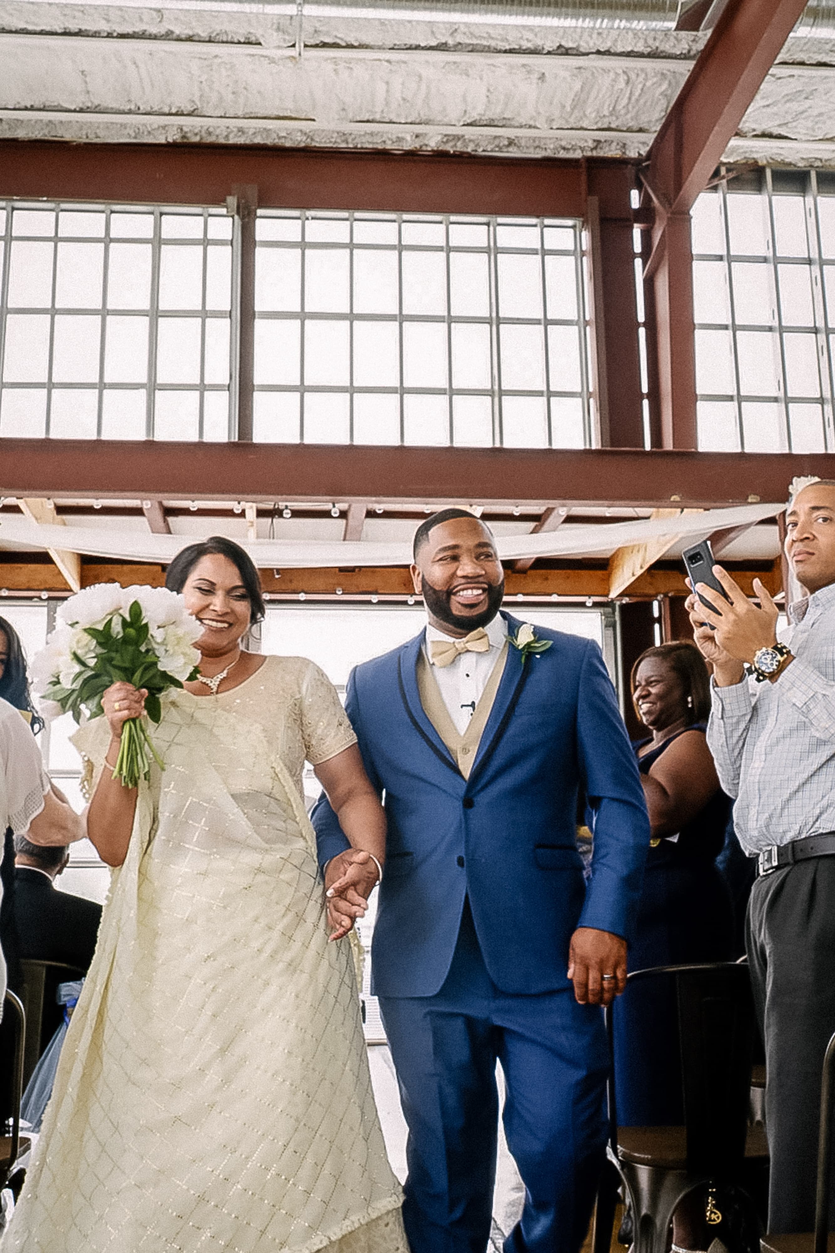 Bride in a cream sari and groom in a blue suit walking back up the aisle smiling in an industrial venue