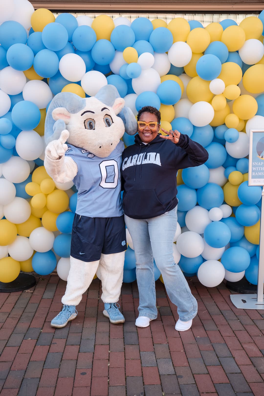 Woman in a Carolina hoodie posing beside the ram mascot in front of a blue, yellow, and white balloon wall