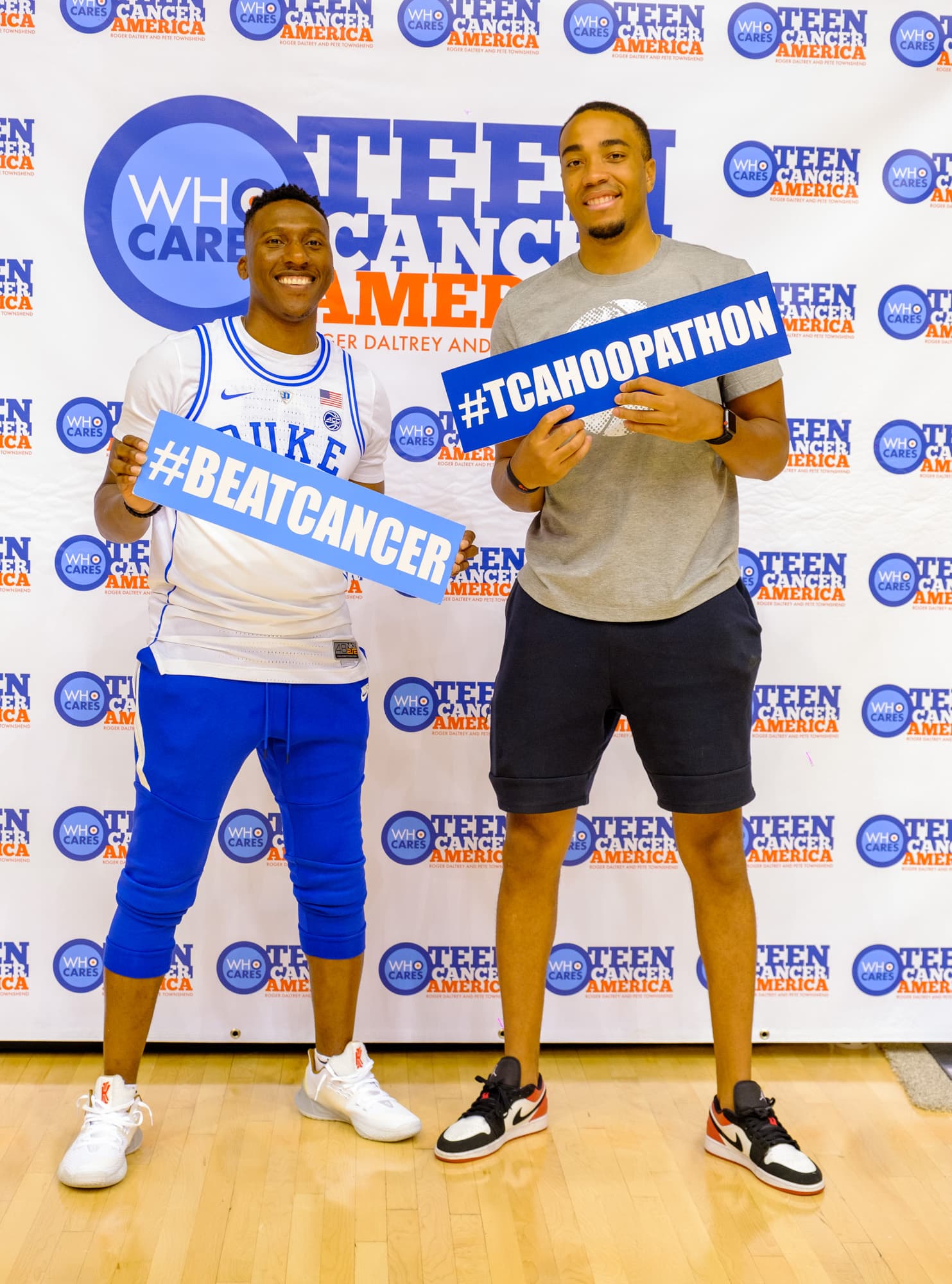 Two men holding Beat Cancer and TCA Hoopathon signs at a Teen Cancer America backdrop