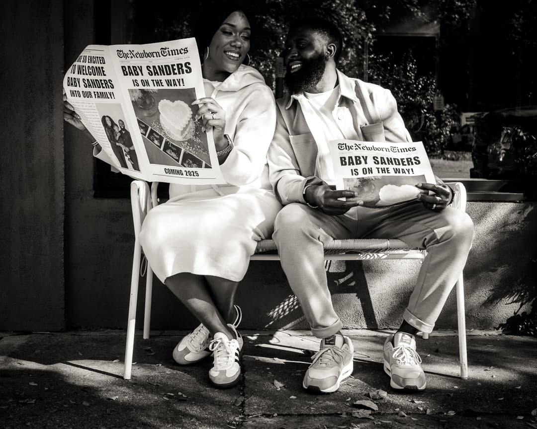 Black-and-white couple seated on a bench holding Baby Sanders Is On The Way newspapers